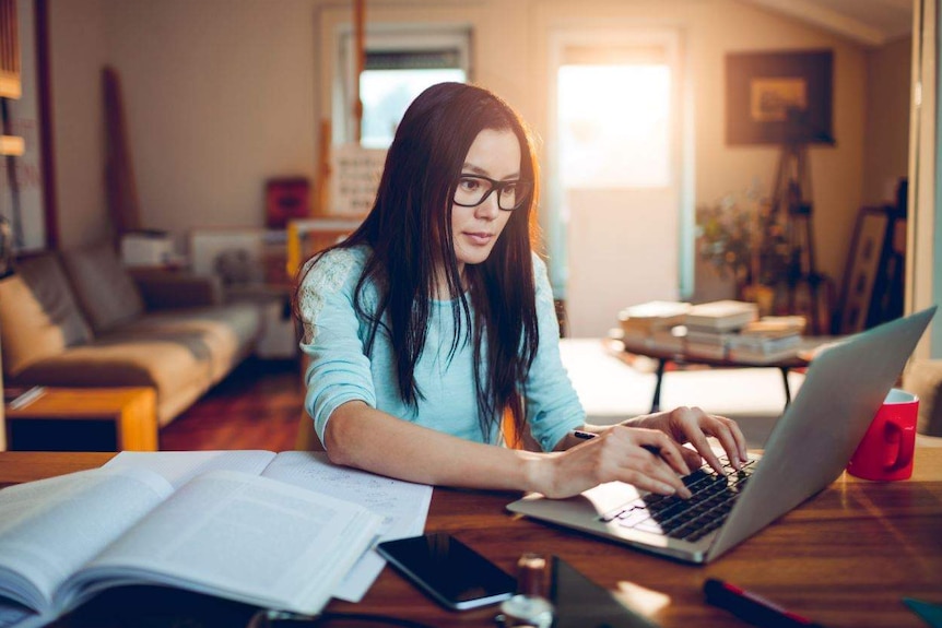 In a dimly lit room a woman sits at a desk covered with open book and phone, looking at a laptop screen.