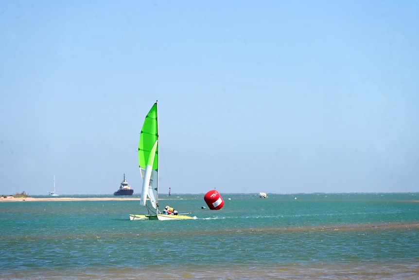 A 14-foot catamaran with a lime green hull and sail maneuvers past a red buoy in a shallow channel. 