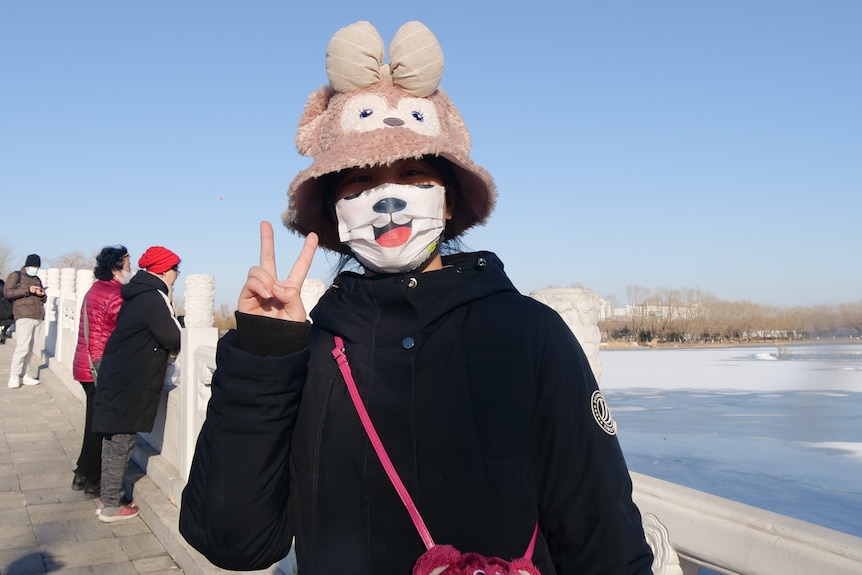 A woman wearing a mask and knitted hat poses with two fingers in the air in a park.