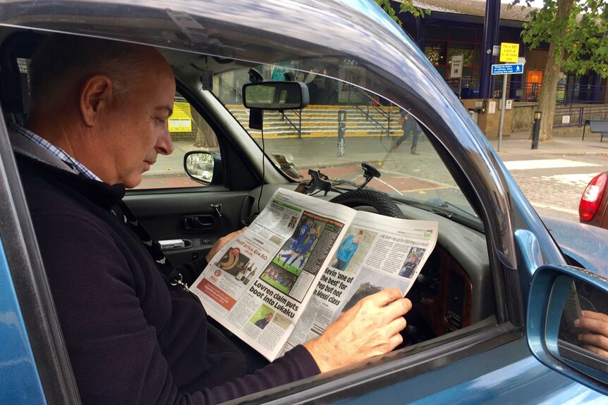 A side-on shot of a taxi driver reading teh newspaper in the driver's seat of his car.