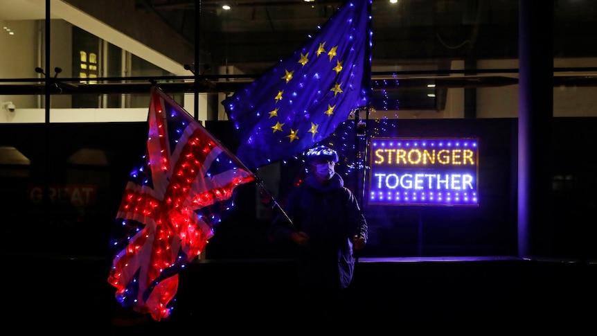 a man holding the Union Jack and EU flag next to a neon sign reading Stronger Together outside a building