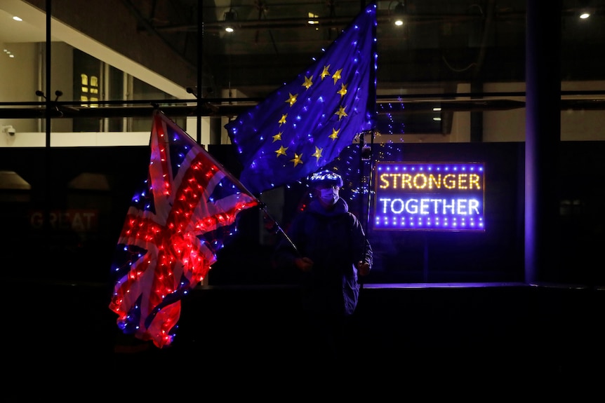 a man holding the Union Jack and EU flag next to a neon sign reading Stronger Together outside a building