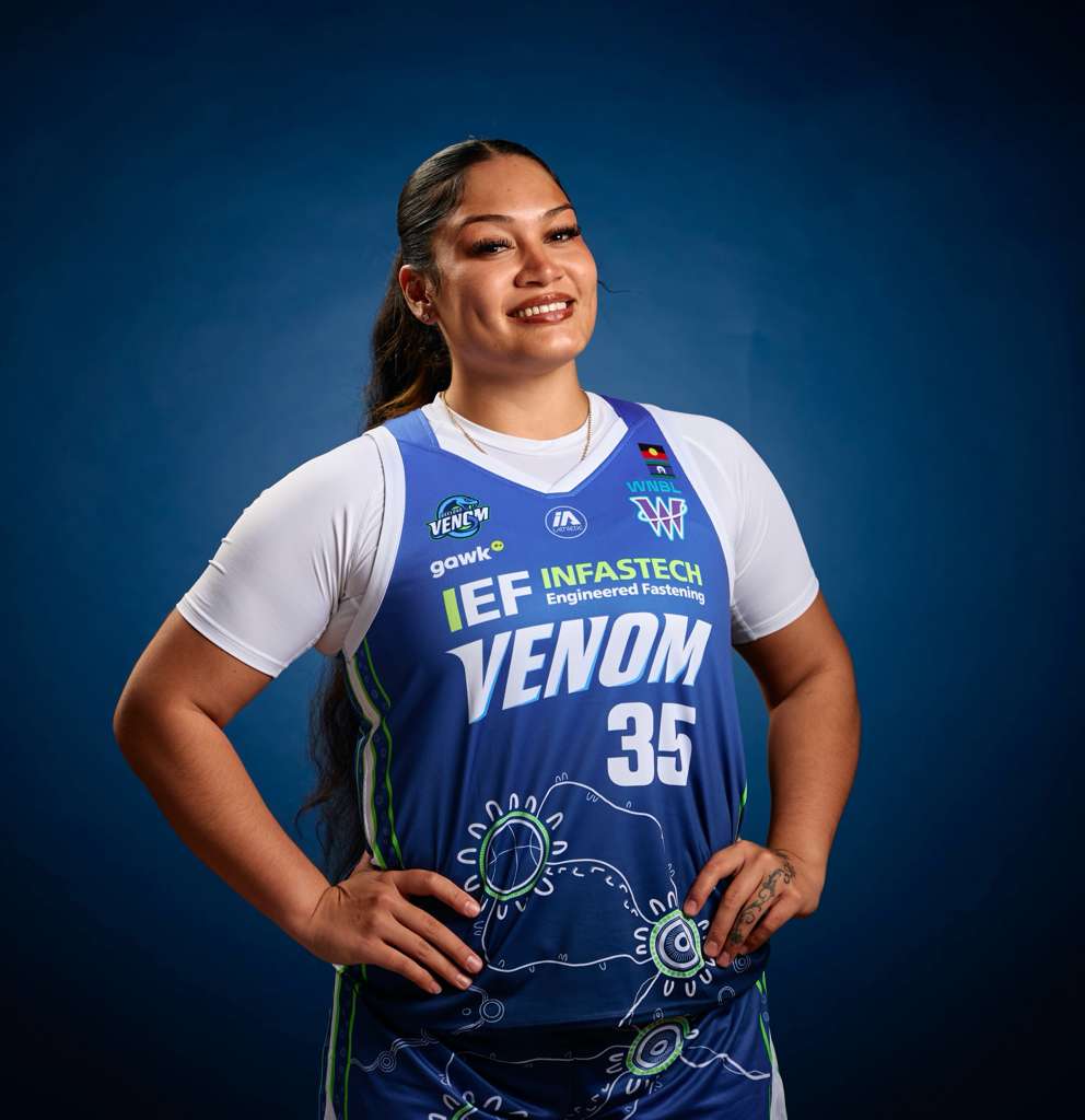 A young lady smiles with her face tilted slightly to the side. she's wearing a white shirt under a purple basketball singlet.