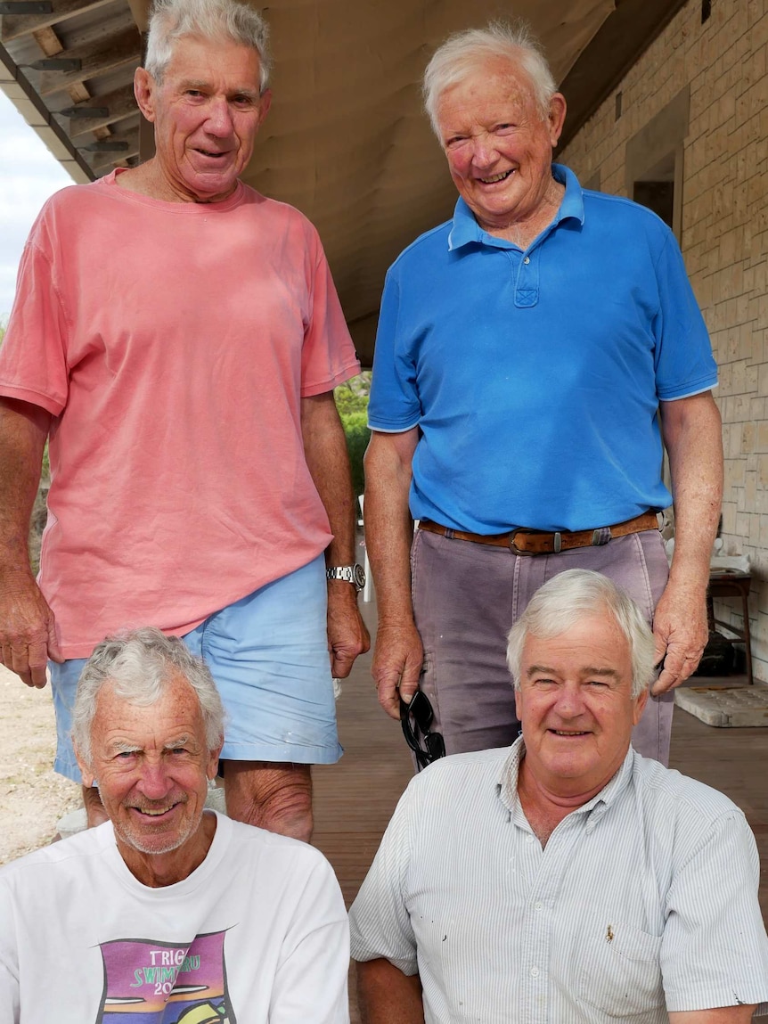 Four white-haired, casually dressed men sit or stand in a group on the verandah of a stone building.