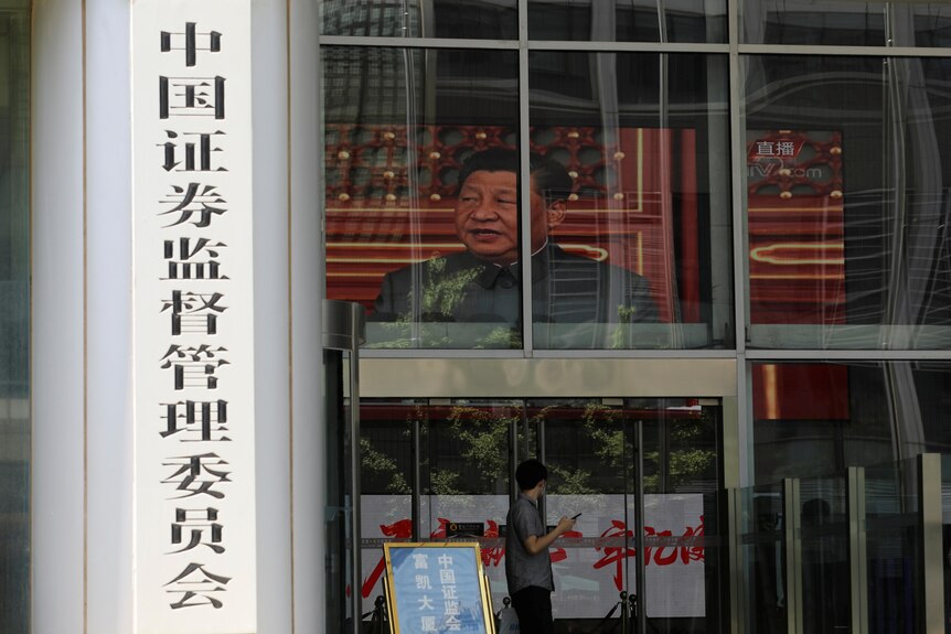 Un homme passe devant un bâtiment avec un panneau indiquant la China Securities Regulatory Commission.  A l'intérieur, le visage de Xi Jinping est sur un écran