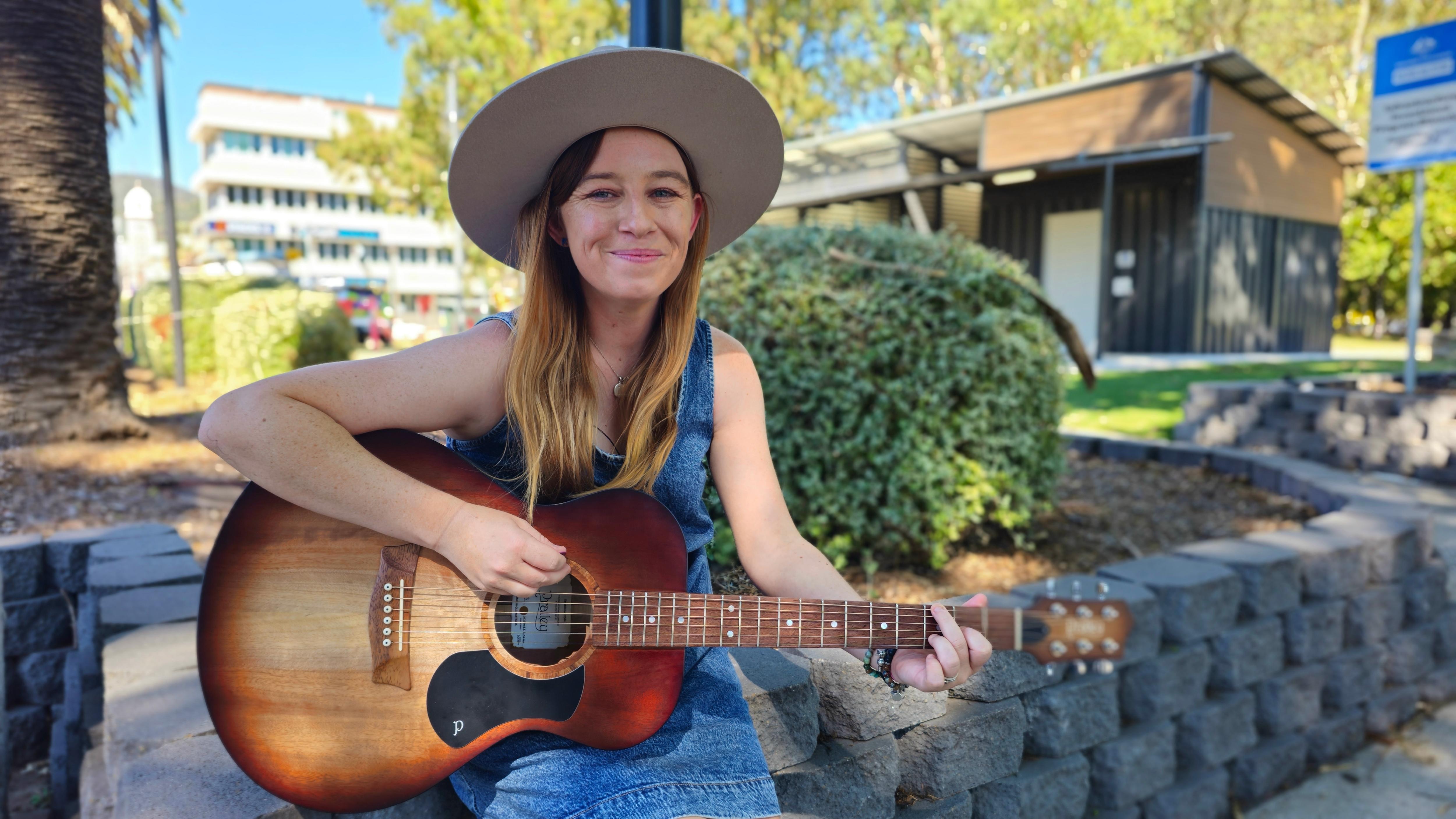 Uma mulher de cabelos compridos, com um vestido azul e um chapéu de abas largas, toca violão em um parque