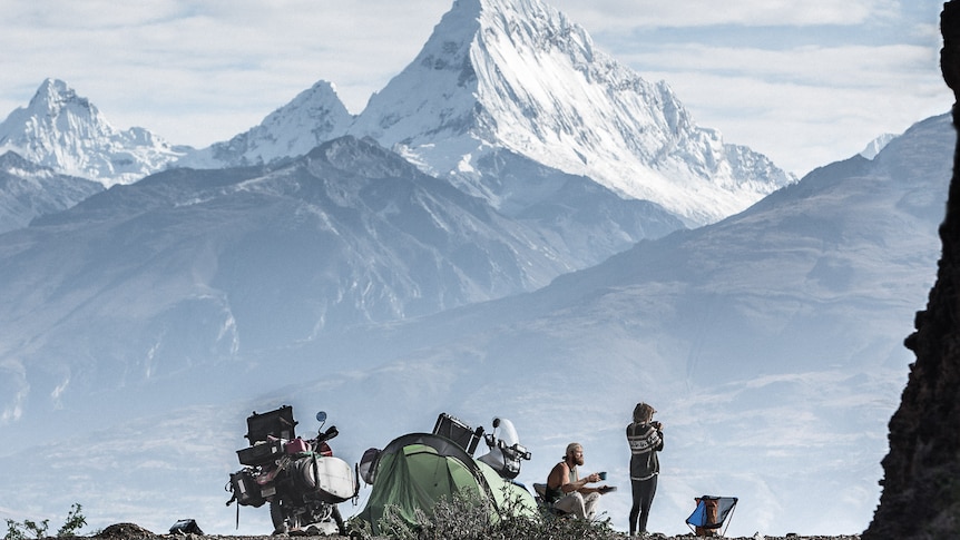A man and a woman are drinking coffee near a tent and bikes with snow-capped mountains in the background