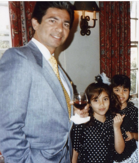 A man in a suit holds a wine glass next to two young girls 