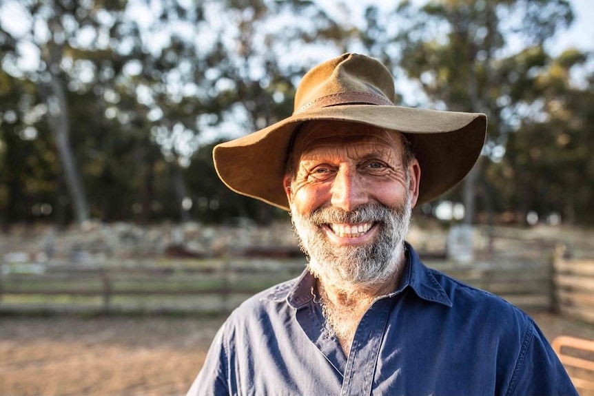 A man wearing a brown hat, blue shirt, smiling with trees in the background.