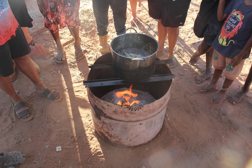 People gathered around a bit metal pot sitting on top of a barrel fire.
