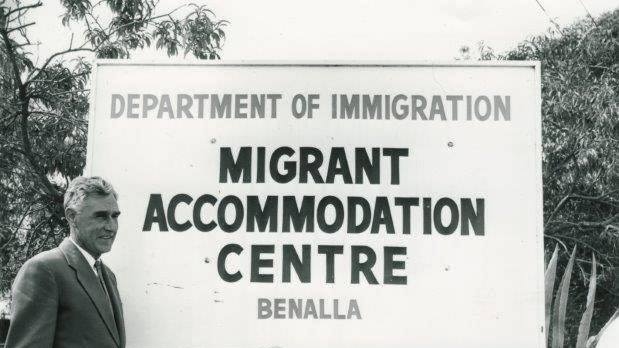 Black and white photo shows a man in a suit lean on a sign that reads 'Department of Immigration Migrant accommodation Benalla'.