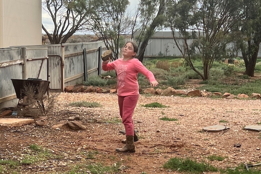 A young girl wearing a pink shirt and pink pants holding a rock in a throwing motion in a back garden.