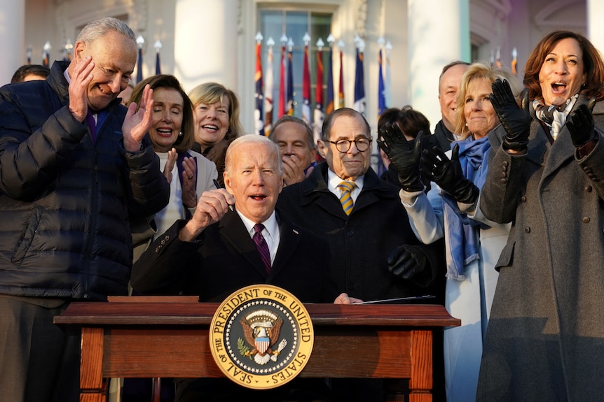 A group of people clapping surrounding Joe Biden sitting at a desk with a pen in his hand