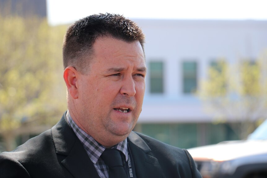 Headshot of a man in a dark suit and check shirt standing outside