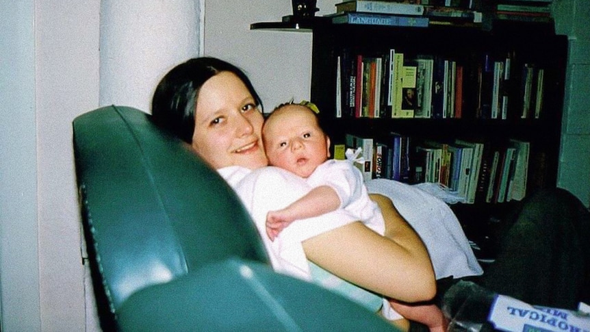 An older photo of a young woman nursing a baby on a leather lounge chair in a living room.
