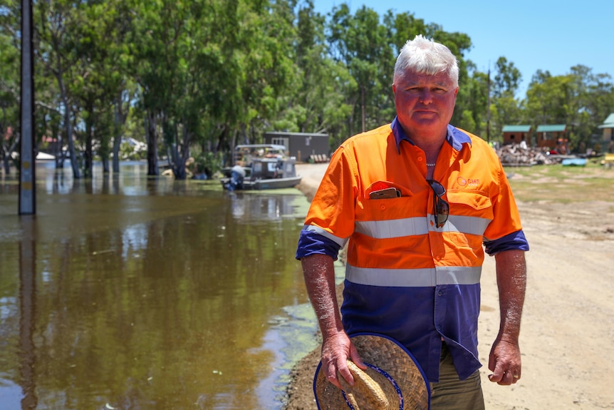 A man wearing an orange and blue shirt with a hat in his hands by his side stands next to water