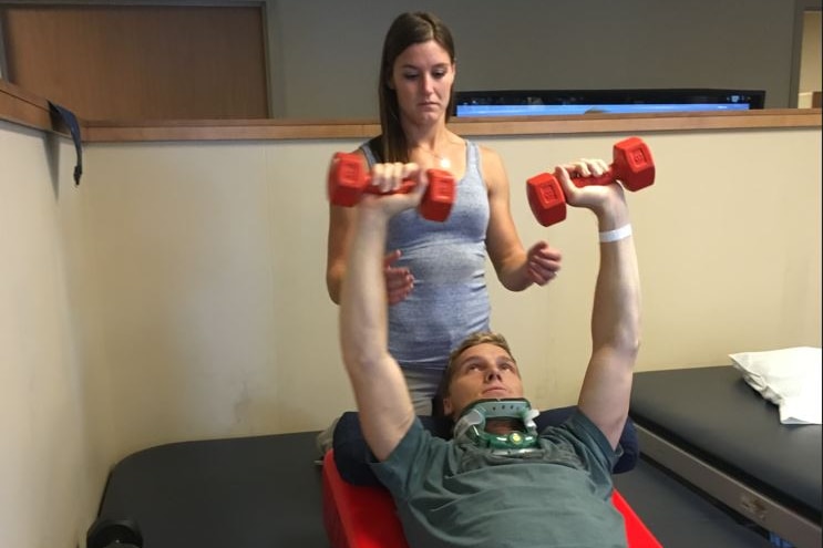 Alise kneels behind Sam Willhoughby as he lifts red dumbellslying on his back