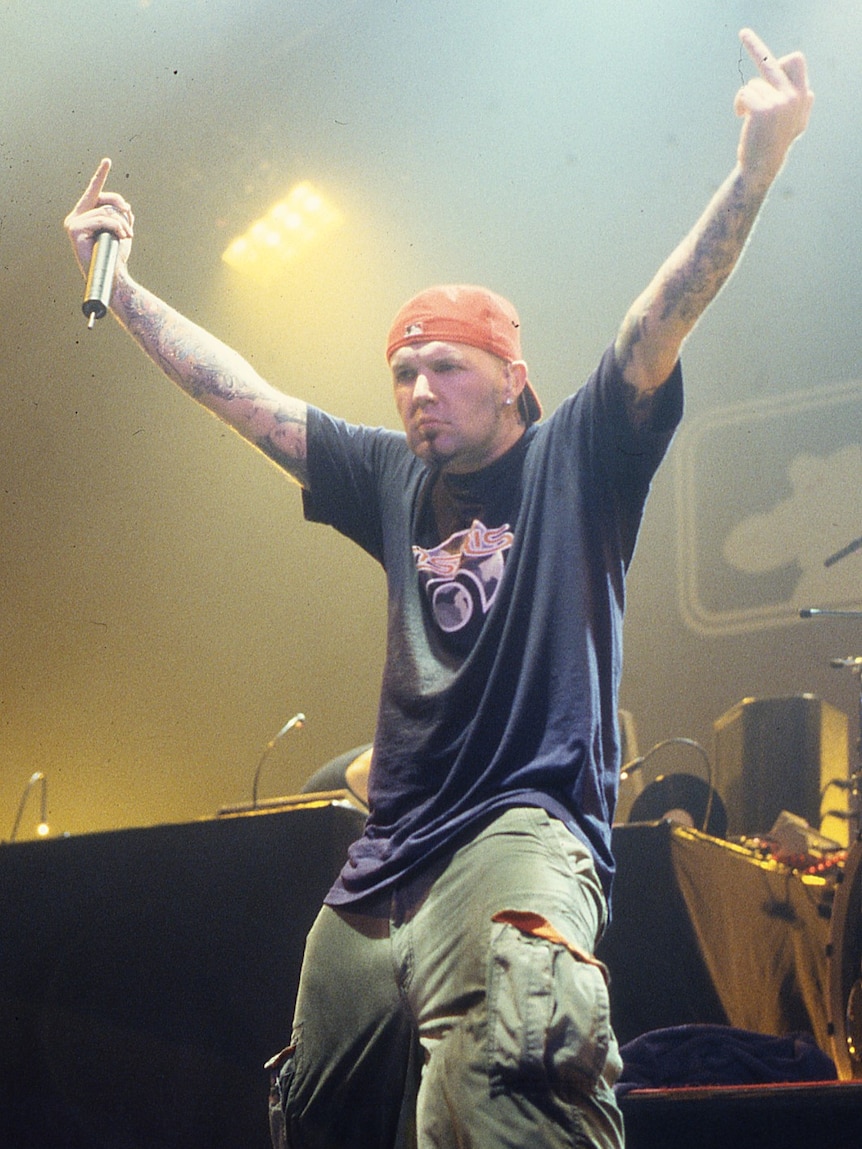 Fred Durst, in red cap and blue t-shirt, gives two extended middle fingers to the crowd while standing on stage