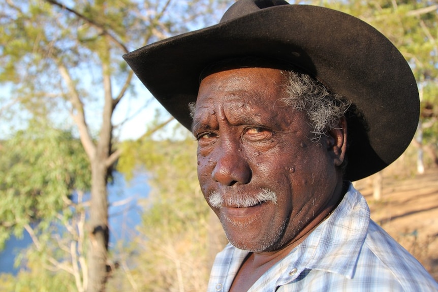 a close up portrait of Frank Shadforth, wearing a hat with trees in the background