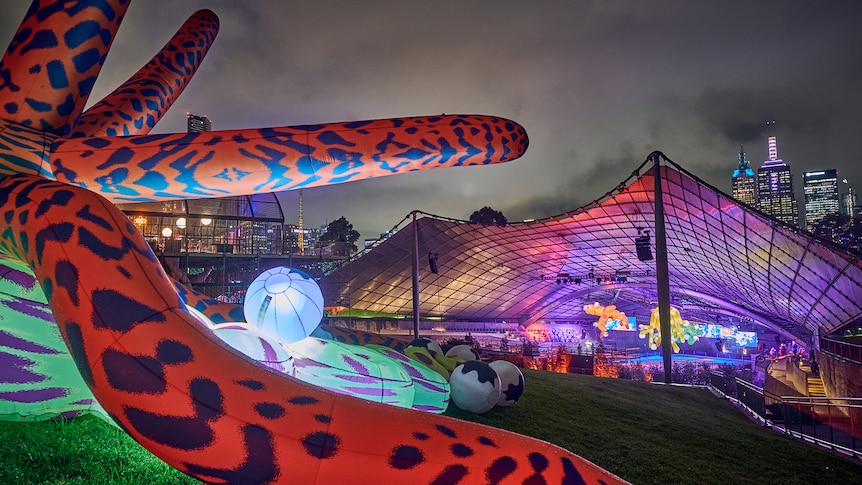 Brightly coloured inflatable sculptures sit atop a grassy hill with a glowing purple ice-skating rink in the background.