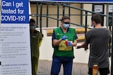 A woman in a green and yellow vest with a face mask and shield uses a red pump near a man in a grey t-shirt.