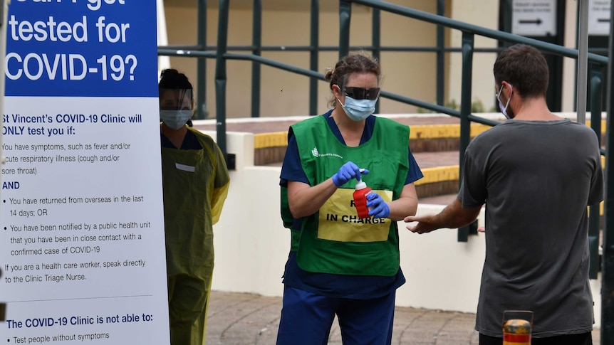 A woman in a green and yellow vest with a face mask and shield uses a red pump near a man in a grey t-shirt.