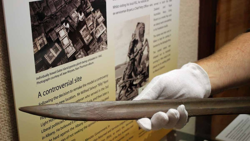 A gloved hand holds a replica bayonet inside a museum cabinet.