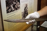 A gloved hand holds a replica bayonet inside a museum cabinet.