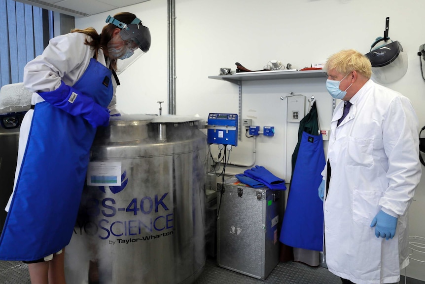 Boris Johnson in a mask looking at a lab worker with her hand in a large storage container.
