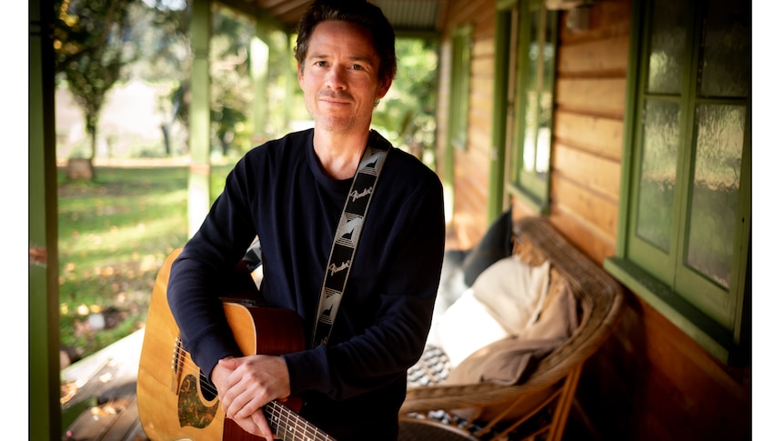 A man holds a guitar outside a country house, he stands on a Verenda and looks directly at the camera. 