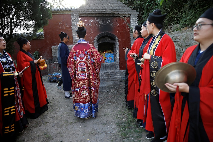 Des personnes en robes de cérémonie, certaines tenant des cymbales, font la queue devant un sanctuaire.