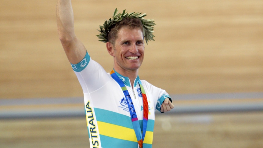 An athlete smiles as he stands on a dais with a gold medal around his neck. 