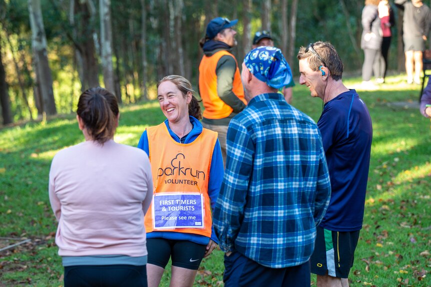 A parkrun volunteer with a first timers and tourists sign stands and smiles.