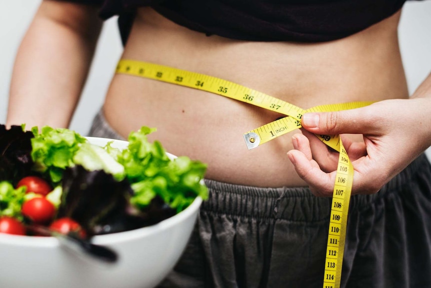 A woman holds a salad in one hand and a tape measure around her belly in the other.