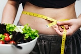 A woman holds a salad in one hand and a tape measure around her belly in the other.