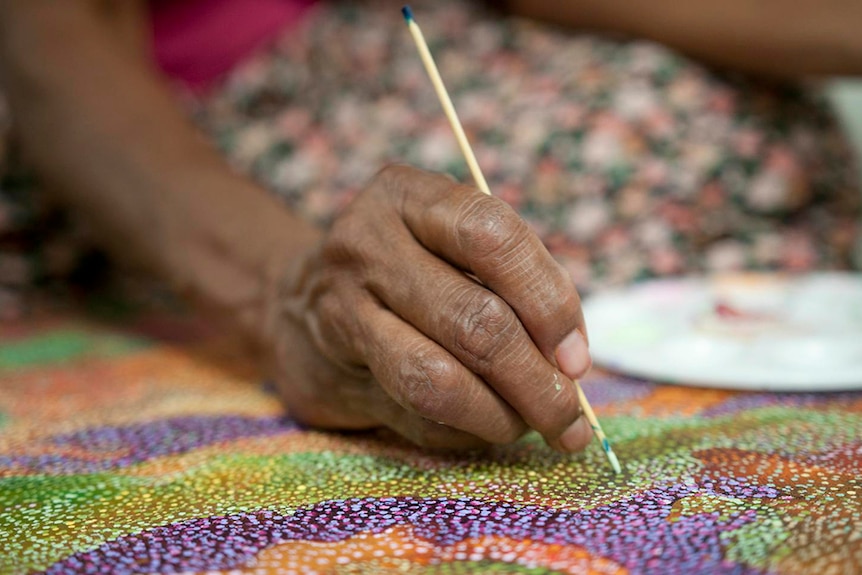 A seated person's hand holds a kebab stick to create a dot shape on colourful dotted painting on canvas.