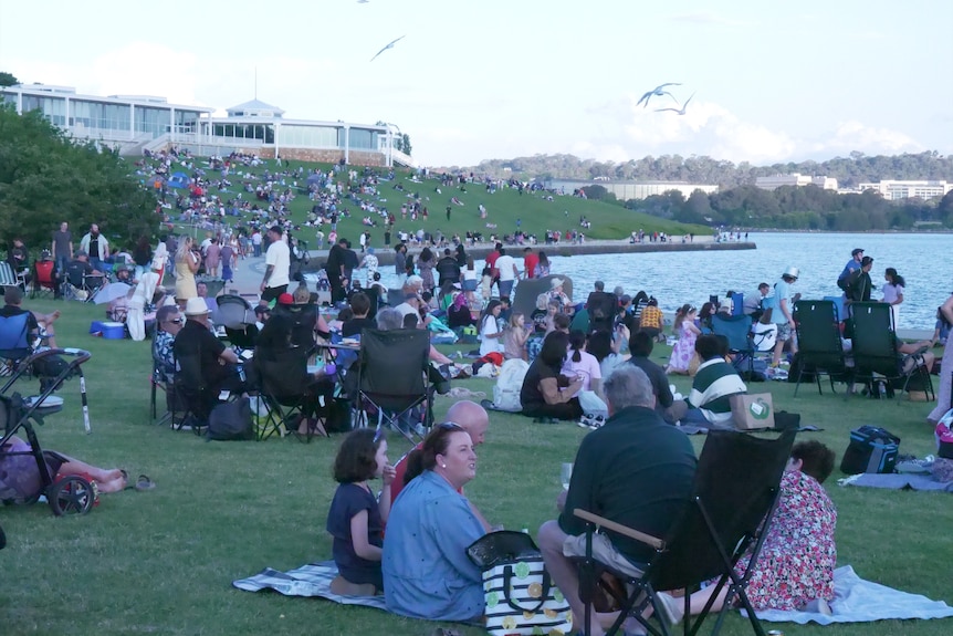 Crowds of people sit on the grassy bank of a lake. 