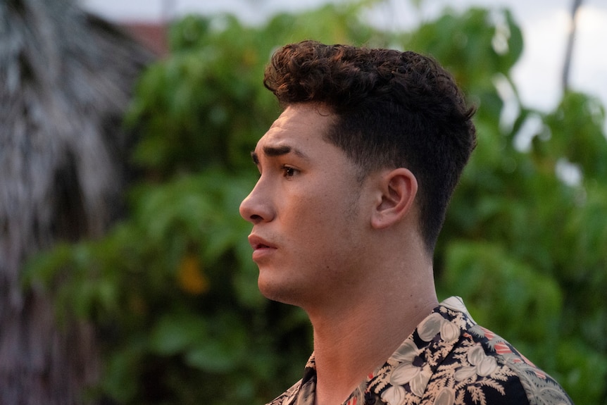 A young Native Hawaiian man looks to the side in concern, tree in the background