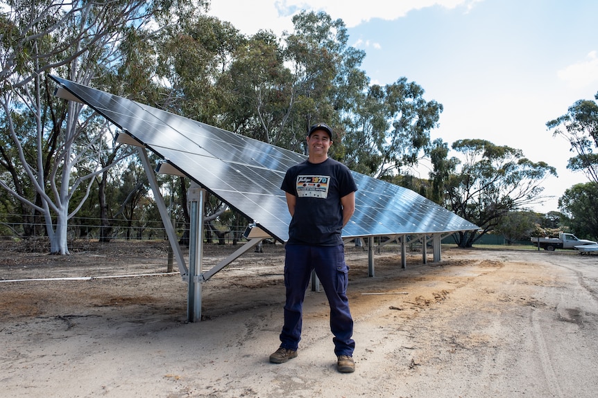 A man in t-shirt and cargo pants stands in front of a ground-mounted solar array