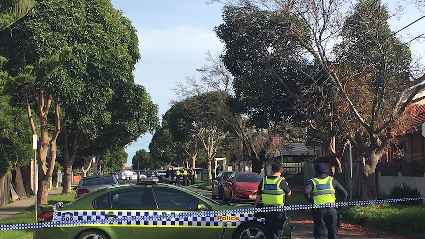 Police stand near a police car blocking off a tree-lined Preston street, while police tape cordons off the area.