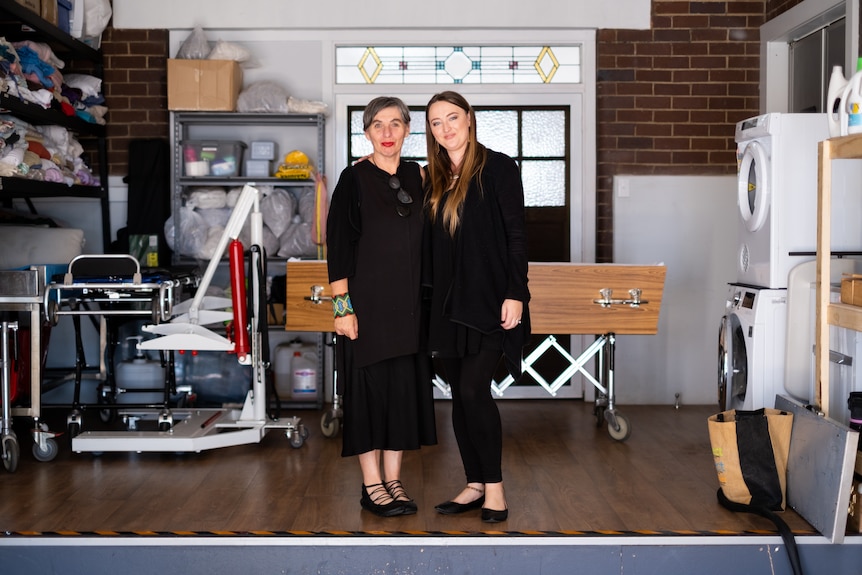 Two women stand in a room that contains two wooden coffins and a washer-dryer, among other items