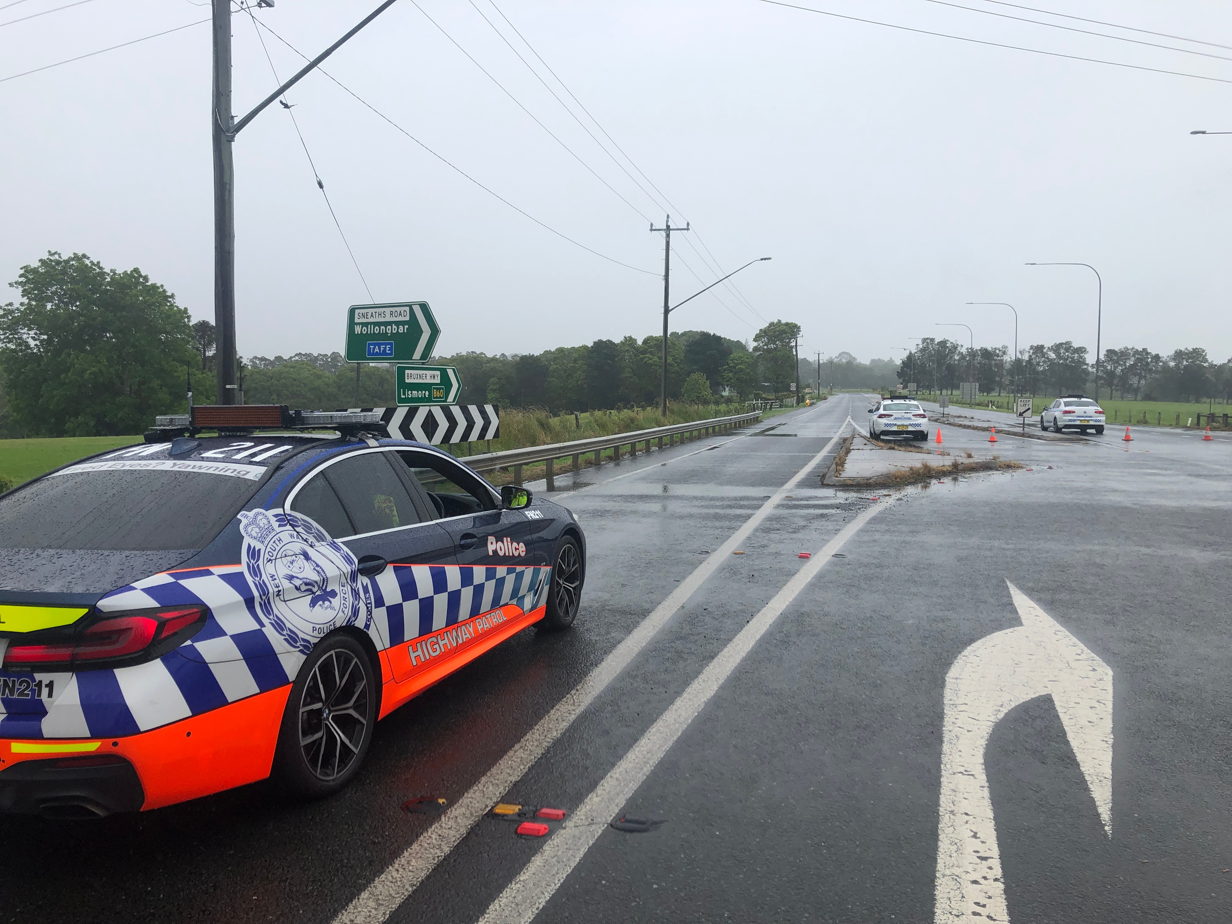 Police cars parked on an empty highway with road signs pointing to Wollongbar and TAFE in the background.