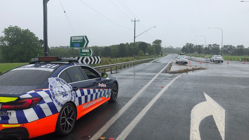 Police cars parked on an empty highway with road signs pointing to Wollongbar and TAFE in the background.
