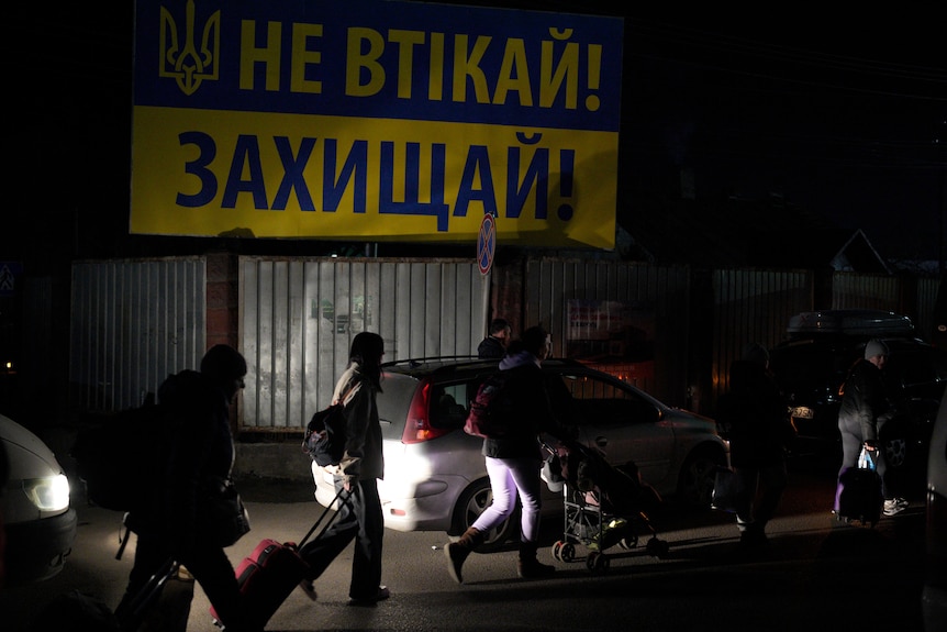 People wearing winter clothes and carrying luggage walk next to a line of cars, with a Ukrainian flag banner on a nearby wall.