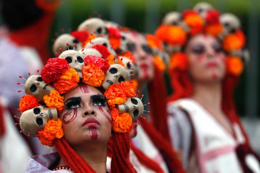 Women in orange wearing flower crowns adorned with skulls look to the sky as they perform