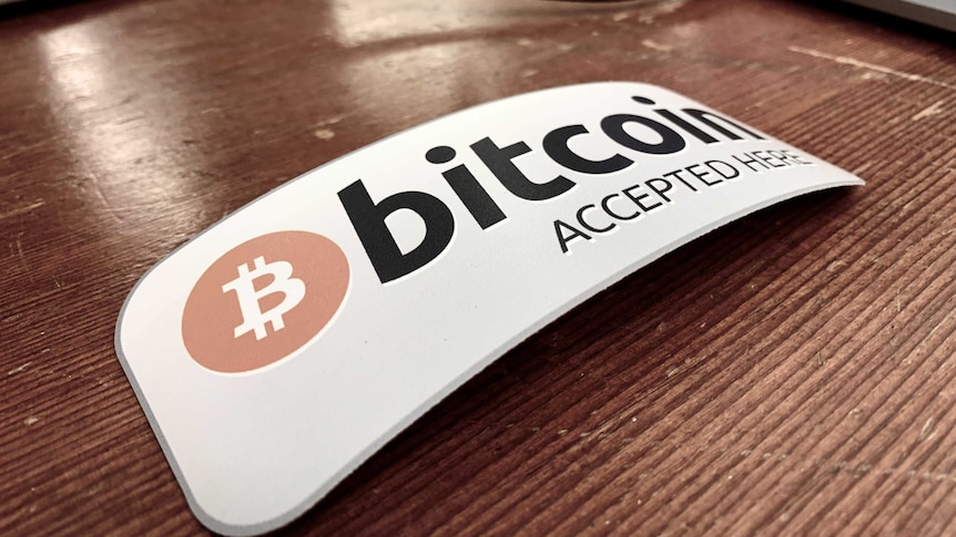 A rectangular sign reading 'bitcoin accepted here' sits on a wooden desk