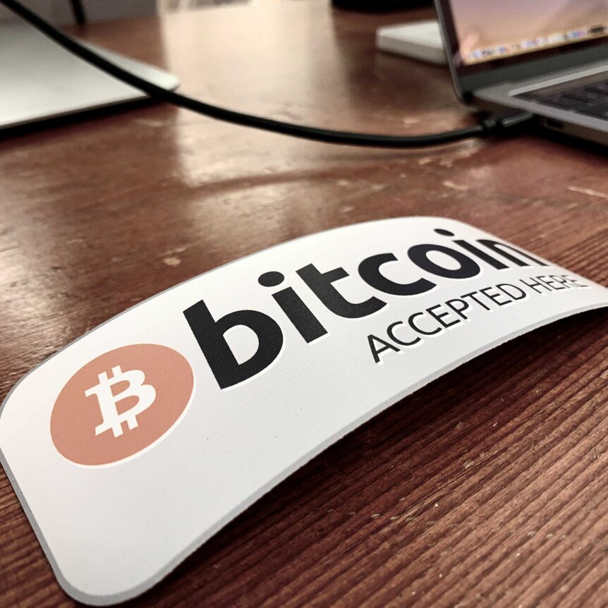 A rectangular sign reading 'bitcoin accepted here' sits on a wooden desk