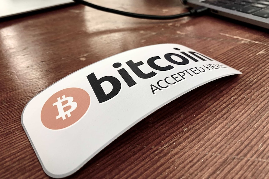 A rectangular sign reading 'bitcoin accepted here' sits on a wooden desk