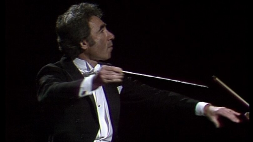 Sergiu Comissiona in profile, dancer-like, with baton raised and left hand outstretched (televised SSO concert, 23 June 1982)