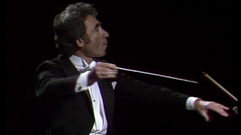 Sergiu Comissiona in profile, dancer-like, with baton raised and left hand outstretched (video still from World of Music recording with Sydney Symphony Orchestra, 23 June 1982)